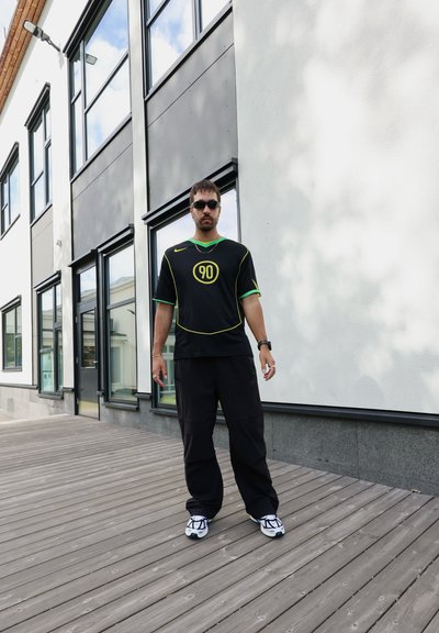 Camiseta deportiva negra con acentos en verde y amarillo, que presenta el número 90. Combinada con pantalones negros holgados y zapatillas deportivas blancas.