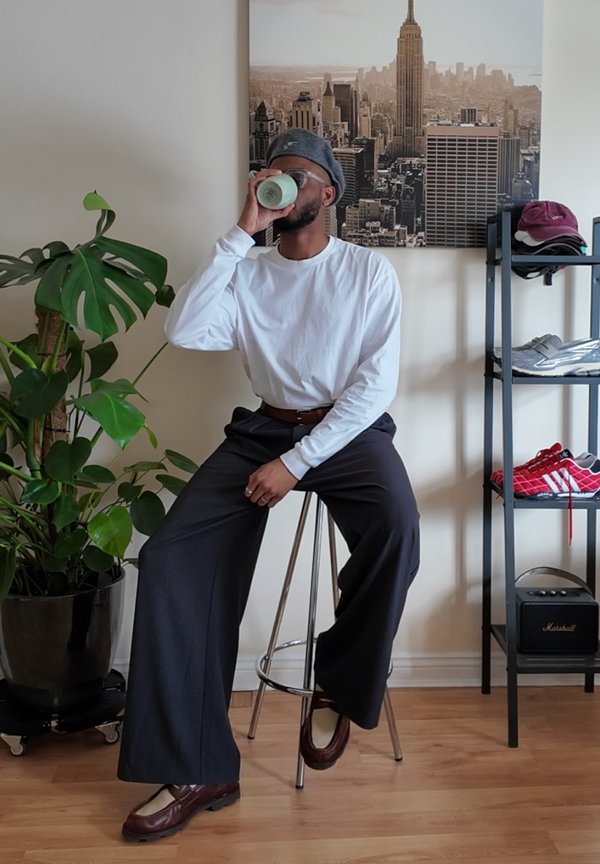 Lässiges Outfit mit einem weißen Langarmshirt, dunklen Hosen und burgunderfarbenen Schuhen. Eine gestrickte graue Mütze setzt Akzente. Der Hintergrund zeigt Grünes.