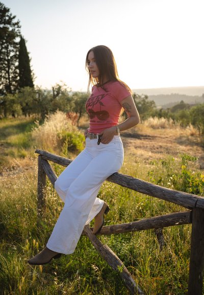 Camiseta rosa ajustada con un gráfico de cereza, pantalones anchos blancos y zapatos marrón claro. De pie junto a una cerca de madera rústica en un campo cubierto de hierba.