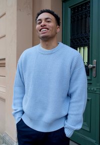 Suéter de malha azul claro com textura canelada, gola redonda e corte descontraído, combinado com calças escuras, em pé perto de uma porta verde.