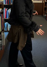 Casaco preto forrado com pele, camisa castanha às quadrados amarrada à cintura, calças pretas e sapatos pontiagudos, com estantes de livros ao fundo.