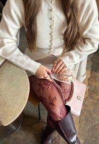 Femme en cardigan crème boutonné, collants en dentelle bordeaux et bottes foncées assise à côté d'une table, tenant un sac à main rose pâle avec un accessoire fleur.
