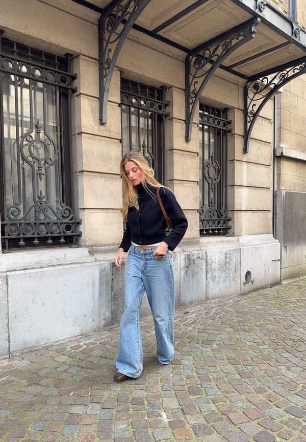 Kvinde med langt blondt hår iført sort sweater og vide jeans går på en brostensbelagt gade ved en stenhøj bygning med ornamenterede vinduer.