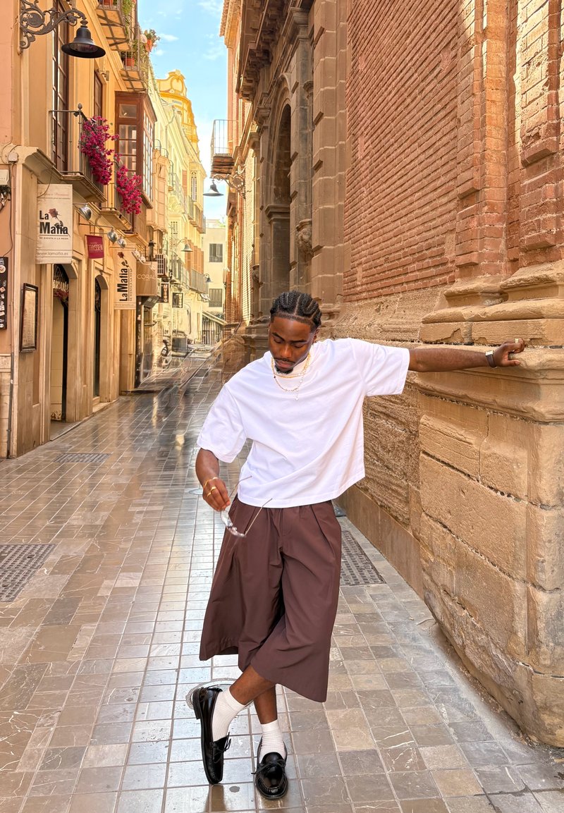 Hombre con camisa blanca y pantalones anchos marrones se apoya en una pared histórica de ladrillo, sosteniendo unas gafas de sol en una calle europea estrecha con tiendas y flores.