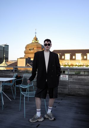 Blazer negro de dos filas de botones sobre una camisa clara, combinado con pantalones cortos negros y zapatillas deportivas gruesas. Los accesorios incluyen gafas de sol oscuras y calcetines a rayas.