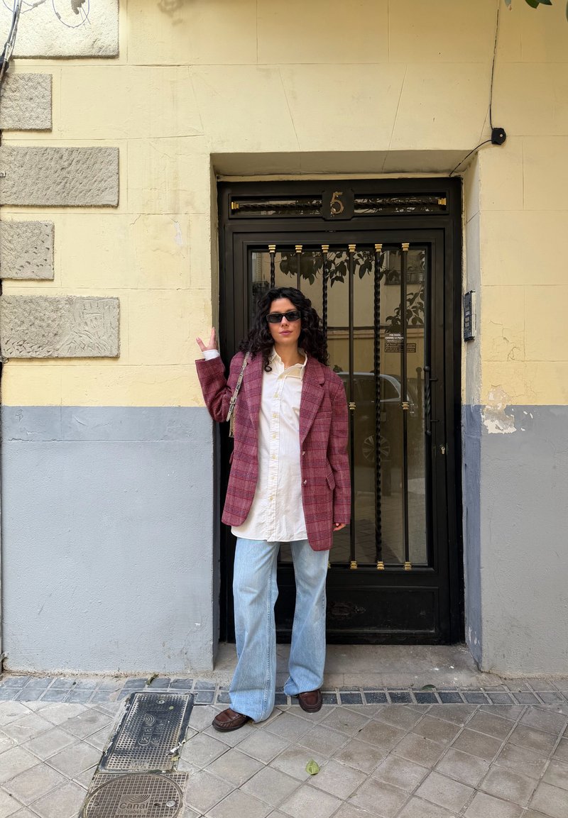 Mujer con cabello rizado que lleva gafas de sol, blazer de cuadros rojos, camisa blanca y jeans azul claro, de pie junto a una puerta metálica negra con el número 5.