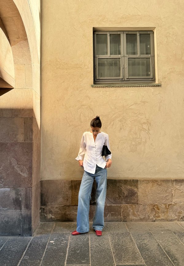 Frau in weißer Bluse, weitem blauen Jeans und roten Schuhen steht und schaut nach unten vor einer beigen Wand mit kleinem Fenster darüber.