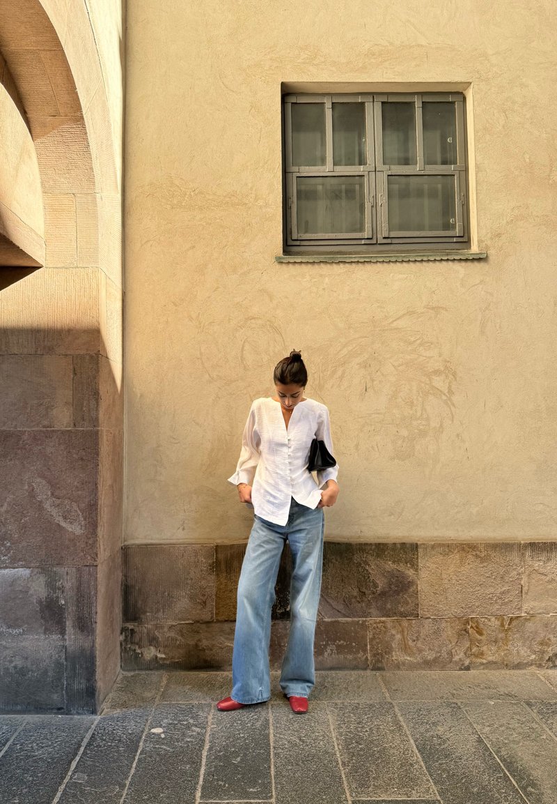 Femme en blouse blanche, jean bleu large et chaussures rouges, debout regardant vers le bas contre un mur beige avec une petite fenêtre au-dessus.