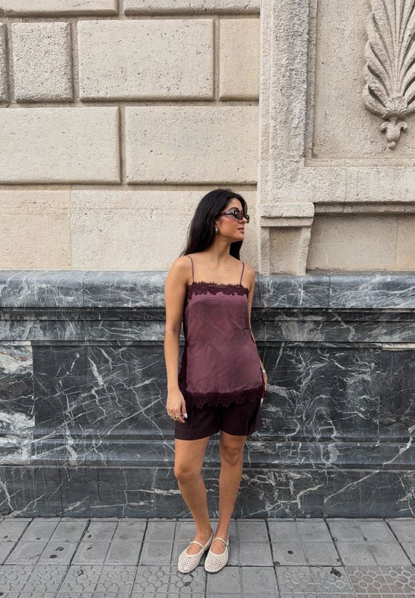 Femme aux longs cheveux foncés porte des lunettes de soleil, un haut violet sans manches, un short sombre et des chaussures blanches tissées, debout contre un mur en pierre.