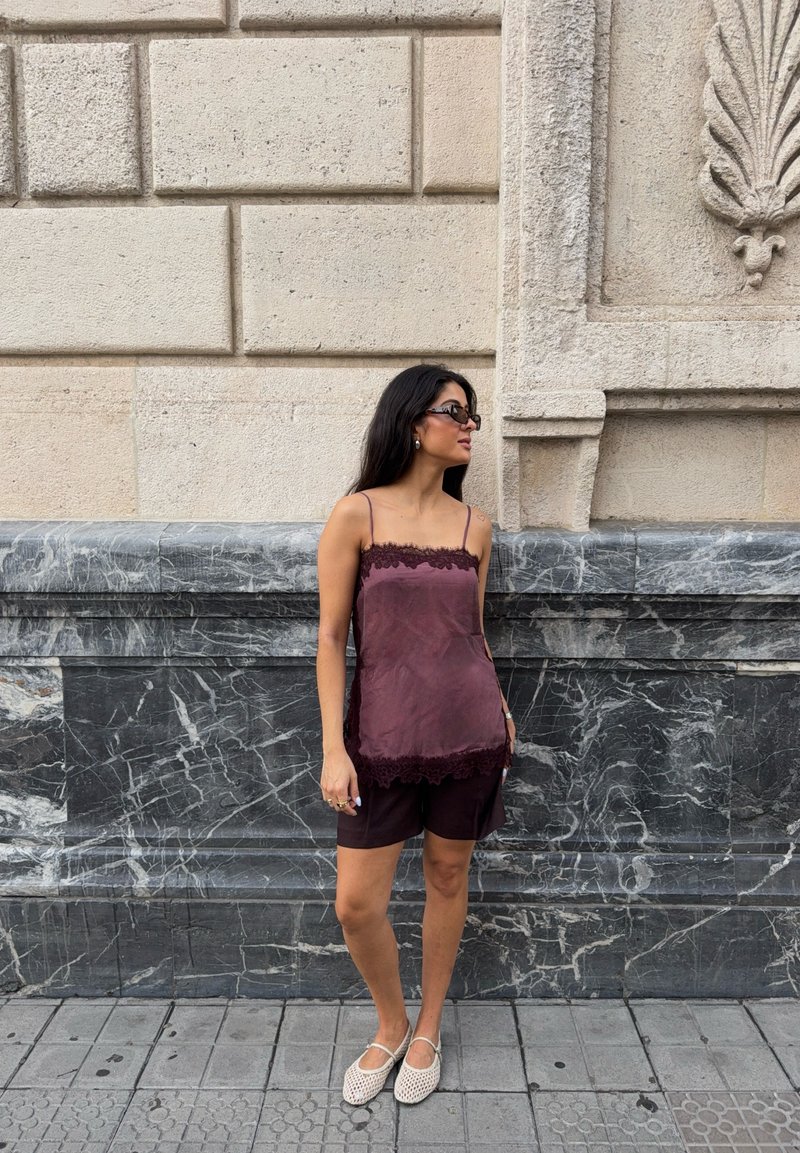 Femme aux longs cheveux foncés porte des lunettes de soleil, un haut violet sans manches, un short sombre et des chaussures blanches tissées, debout contre un mur en pierre.