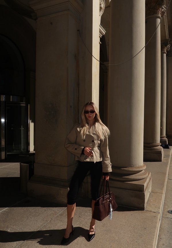 Casaco trinchado curto bege, leggings pretas, saltos pretos. Bolsa castanha com um charme. O fundo apresenta colunas clássicas. Sombras da luz solar.