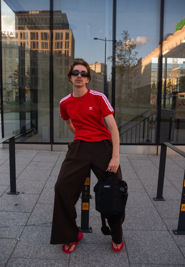 Persona joven con gafas de sol vistiendo una camiseta roja de Adidas y pantalones anchos negros, sosteniendo una bolsa negra, posando al aire libre junto a paredes de cristal.