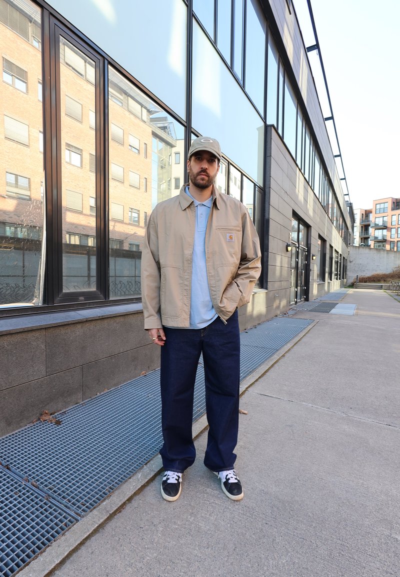 Bărbat purtând o jachetă bej, cămașă albastră, pantaloni închisi la culoare și șapcă, stă pe trotuar lângă o clădire din sticlă și cărămidă într-un mediu urban.