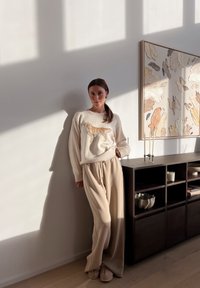 Bežový sveter s dizajnom zlatého retrievera, krémové nohavice s širokými nohavicami a mäkké papuče. Pozadie obsahuje minimalistickú poličku a umelecké dielo.