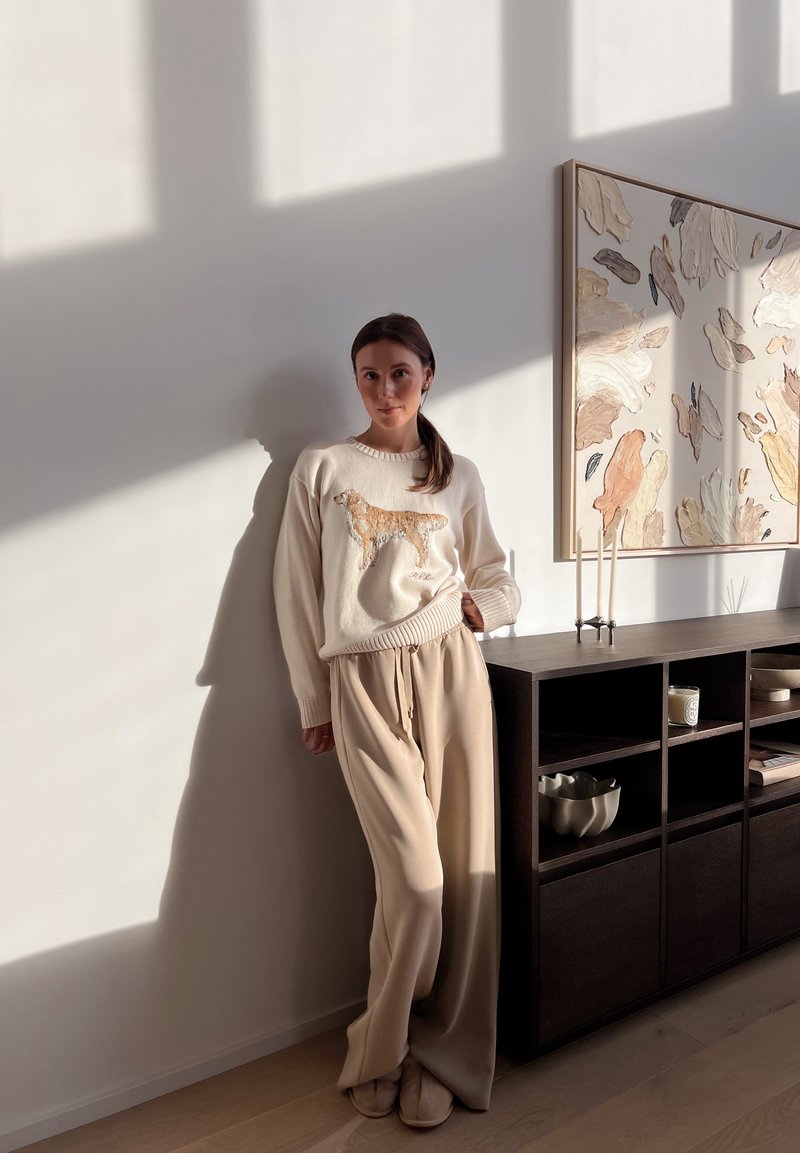 Beige jumper featuring a golden retriever design, cream wide-leg trousers, and soft slippers. The background includes a minimalist shelf and artwork.