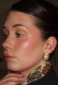 Jeune femme à la peau lisse, avec quelques taches de rousseur légères, piercings au nez et à l'oreille, portant une grande boucle d'oreille en perle, et une écharpe à motif floral, touchant son menton.