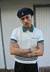 Λευκό πλεκτό γιλέκο με πράσινο γιακά πάνω από πουκάμισο με γιακά, συνδυασμένο με μαύρη μπέρα και μαύρο αθλητικό παντελόνι. Τατουάζ ορατά στα χέρια.