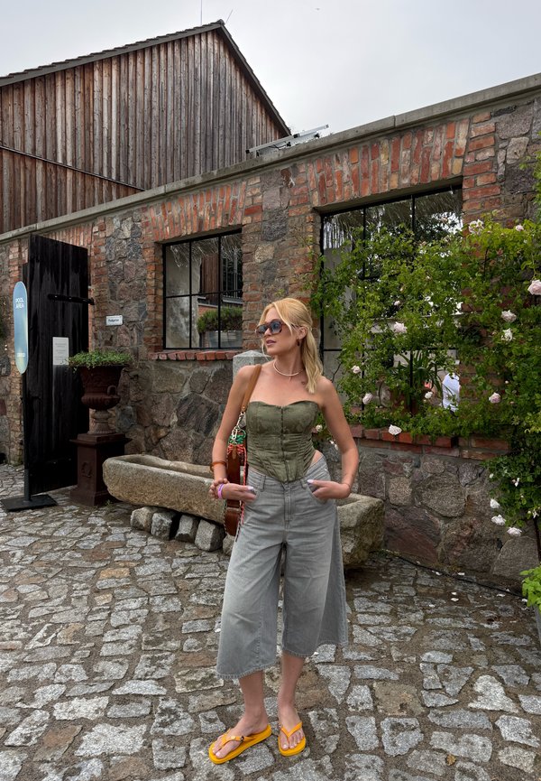 Vrouw met blond haar, die een zonnebril, een groene strapless top, grijze wijde cropped broek en gele sandalen draagt, staat op de kasseien bij een bakstenen gebouw.