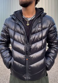 Personne barbu portant une veste matelassée noire brillante sur un sweat à capuche noir et deux colliers à chaîne, dont un avec un pendentif ancre, à l'extérieur.