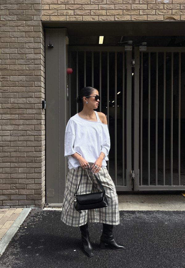 Vrouw draagt een witte off-shoulder top, geruite wijde broek, zwarte laarzen en zonnebril, staat bij een omheinde ingang en houdt een zwarte handtas vast.