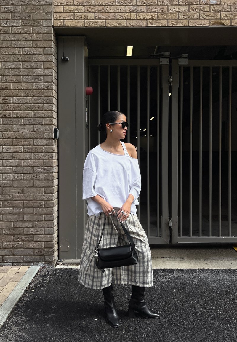 Kvinde iført hvid off-shoulder top, ternede vidde bukser, sorte støvler og solbriller står ved en indhegnet indgang og holder en sort håndtaske.