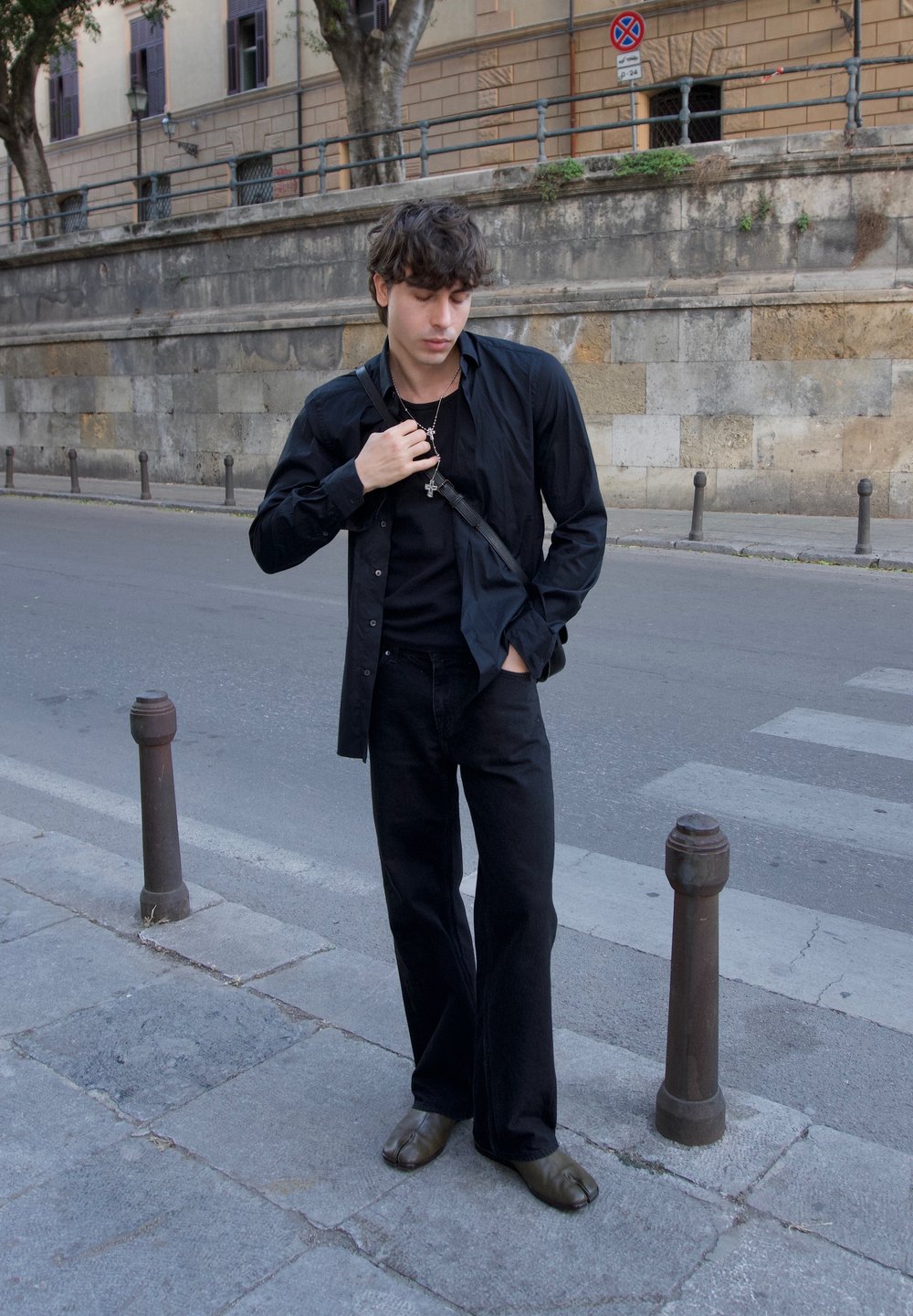 Fekete hosszúujjú ing egy fekete póló felett, széles lábú fekete nadrággal és barna slip-on cipővel, egy utcán állva.