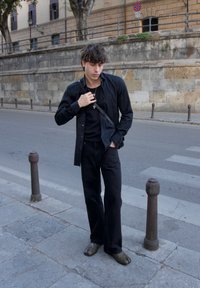 Fekete hosszúujjú ing egy fekete póló felett, széles lábú fekete nadrággal és barna slip-on cipővel, egy utcán állva.