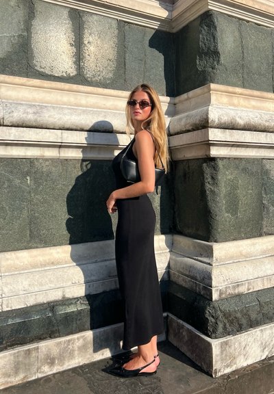 Vestido maxi negro ajustado, bolso negro y bailarinas negras de punta. De pie contra una pared de piedra gris texturizada con acentos de mármol blanco.