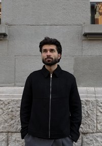 Homme barbu portant une veste noire zippée, debout contre un mur en pierre, regardant légèrement vers le haut avec les mains dans les poches.