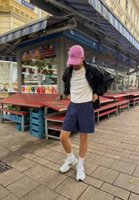 Svart zip-up jacka, vit T-shirt, marinblå shorts, rosa keps med "GAP", vita sneakers med gröna detaljer, står nära matstånd på kullersten.