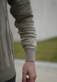 Manche de pull en maille beige avec des détails texturés ondulés et un poignet côtelé. Le tissu semble doux avec une coupe ample. Variations subtiles de couleur.