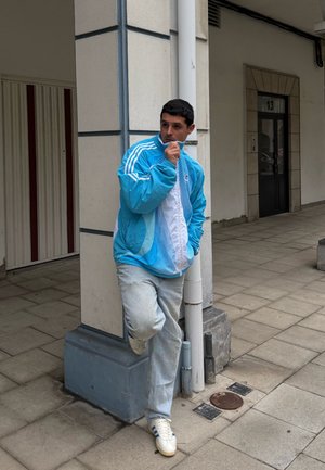 Chaqueta deportiva azul y blanca con cremallera y rayas, jeans azul claro y zapatillas blancas. De pie contra una columna en una zona de azulejos.