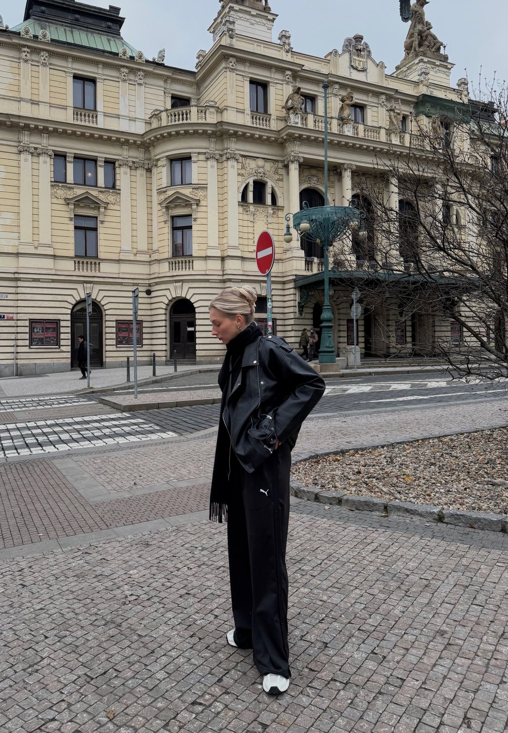 Femme en veste et pantalon en cuir noir debout sur une rue pavée près d'un bâtiment historique beige avec une façade ornée et des panneaux de signalisation.