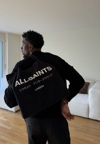 Homme en pull noir portant un sac fourre-tout noir avec l'inscription "ALLSAINTS LONDON" et du texte japonais, debout dans un salon moderne avec un sol en bois.