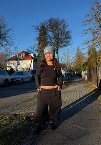 Jeune femme en crop top noir, pantalon ample et bonnet gris, debout sur un trottoir ensoleillé dans une zone résidentielle avec des arbres dépouillés et des voitures garées.