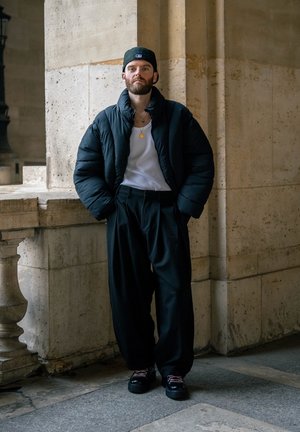 Hombre con barba, vestido con chaqueta negra acolchada, camisa blanca, pantalones negros holgados y gorra negra puesta al revés, de pie dentro de un edificio con columnas de piedra.