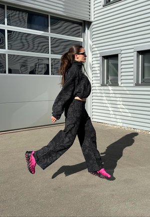 Fekete virágmintás oversized pulóver és hozzáillő bőszárú nadrág, élénk rózsaszín cipővel, texturált talppal, mozgás közben.