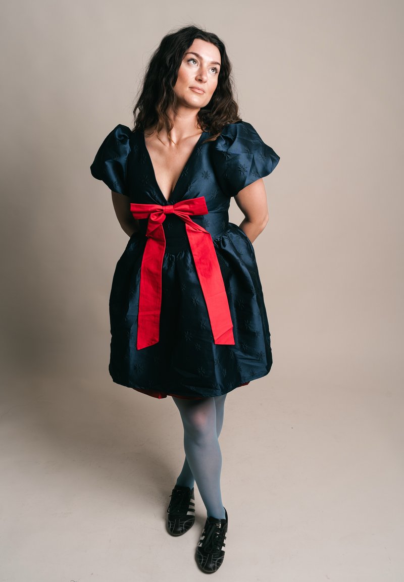 Göndör sötét hajú nő fekete ruhában, mely puffadt ujjakkal és nagy piros masnival díszített, szürke harisnyában és fekete cipőben, kezeit a háta mögött tartva áll.