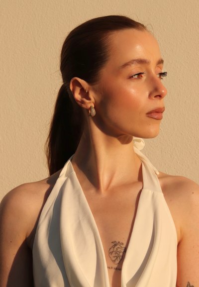 Top blanco tipo halter con suaves drapeados y textura lisa. La modelo tiene el cabello largo y castaño, lleva pendientes de oro y plata, y un tatuaje en forma de corazón visible en el pecho.