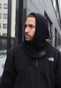 Czarna dzianinowa kominiarka o prążkowanej fakturze, noszona na czarnej kurtce zapinanej na zamek błyskawiczny z logo. Na tle nowoczesnego, miejskiego krajobrazu.