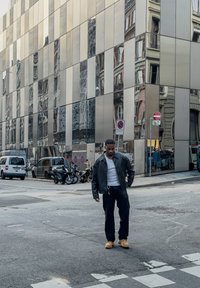 Peegeldav metallist hoone fassaad erinevates toonides; tänaval seisab musta jope ja kollaste kingadega inimene, lähedal on sõidukid.