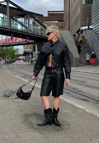 Chaqueta corta de cuero negro con gran cuello, pantalones cortos a juego y botas blancas. Los accesorios incluyen un bolso negro con correa de cadena.