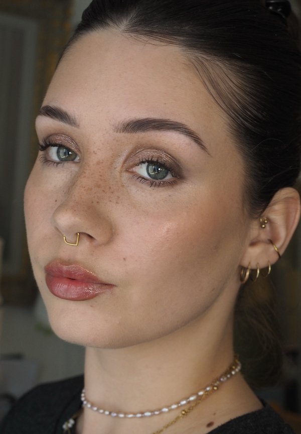 Jonge vrouw met groene ogen, sproeten, gouden septumring, meerdere gouden oorbellen, parelketting en subtiele make-up die iets opzij kijkt.