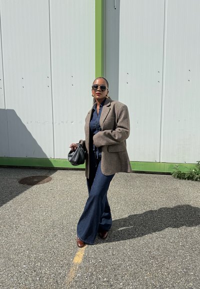 Blazer de lana marrón sobre una camisa de denim oscuro, combinado con pantalones de denim acampanados y zapatos marrones, accesorios con un bolso negro y gafas de sol.