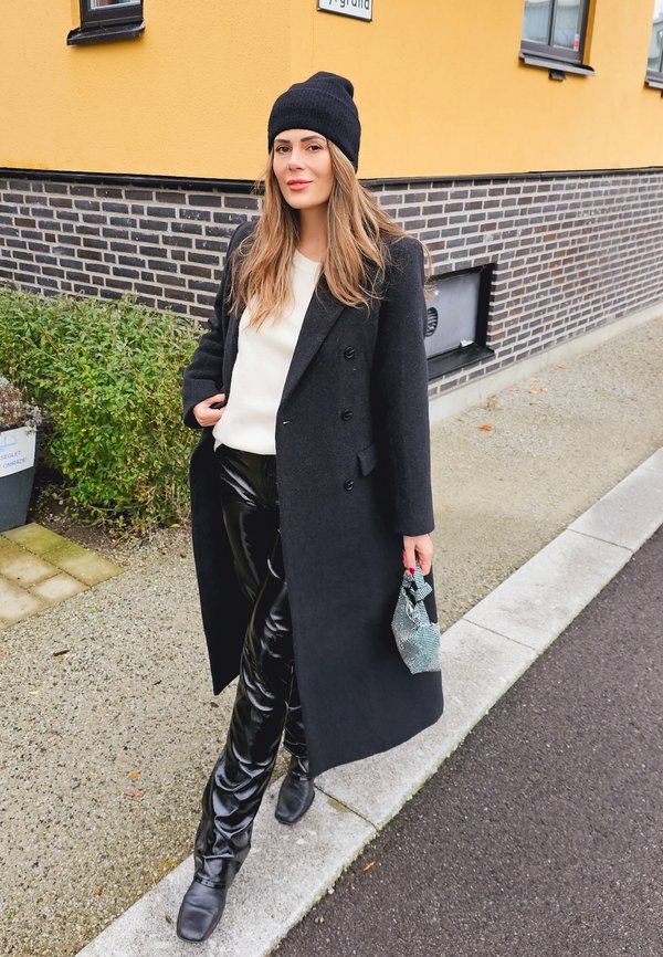 Lungo cappotto nero su un maglione bianco, pantaloni neri lucidi e stivali neri. Indossa un berretto nero e tiene in mano una piccola borsa con motivo.