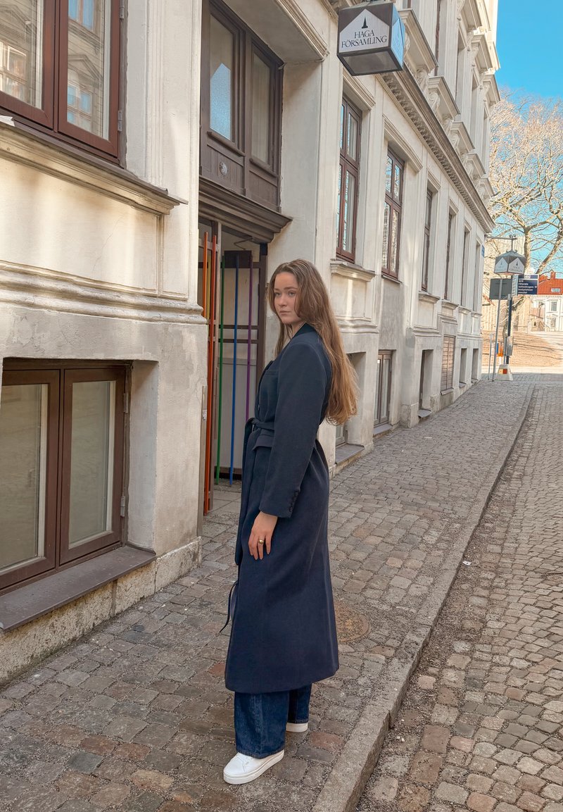 Femme en long manteau sombre et baskets blanches, debout sur une rue pavée près d'une entrée de bâtiment avec une enseigne "Haga Församling".