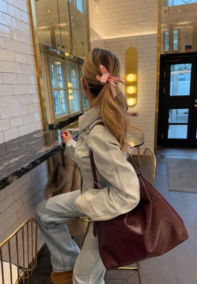 Chaqueta de denim y jeans azul claro, botas marrones, gran bolso de mimbre burdeos, cinta rosa en el cabello, encimera de mármol, acentos dorados en el fondo.