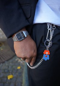 Srebrni sat s pravokutnim okvirom i prugastim remenom, pričvršćen na crni pojas s crvenim i plavim cvjetnim privjescima. Crne hlače i džep su vidljivi.