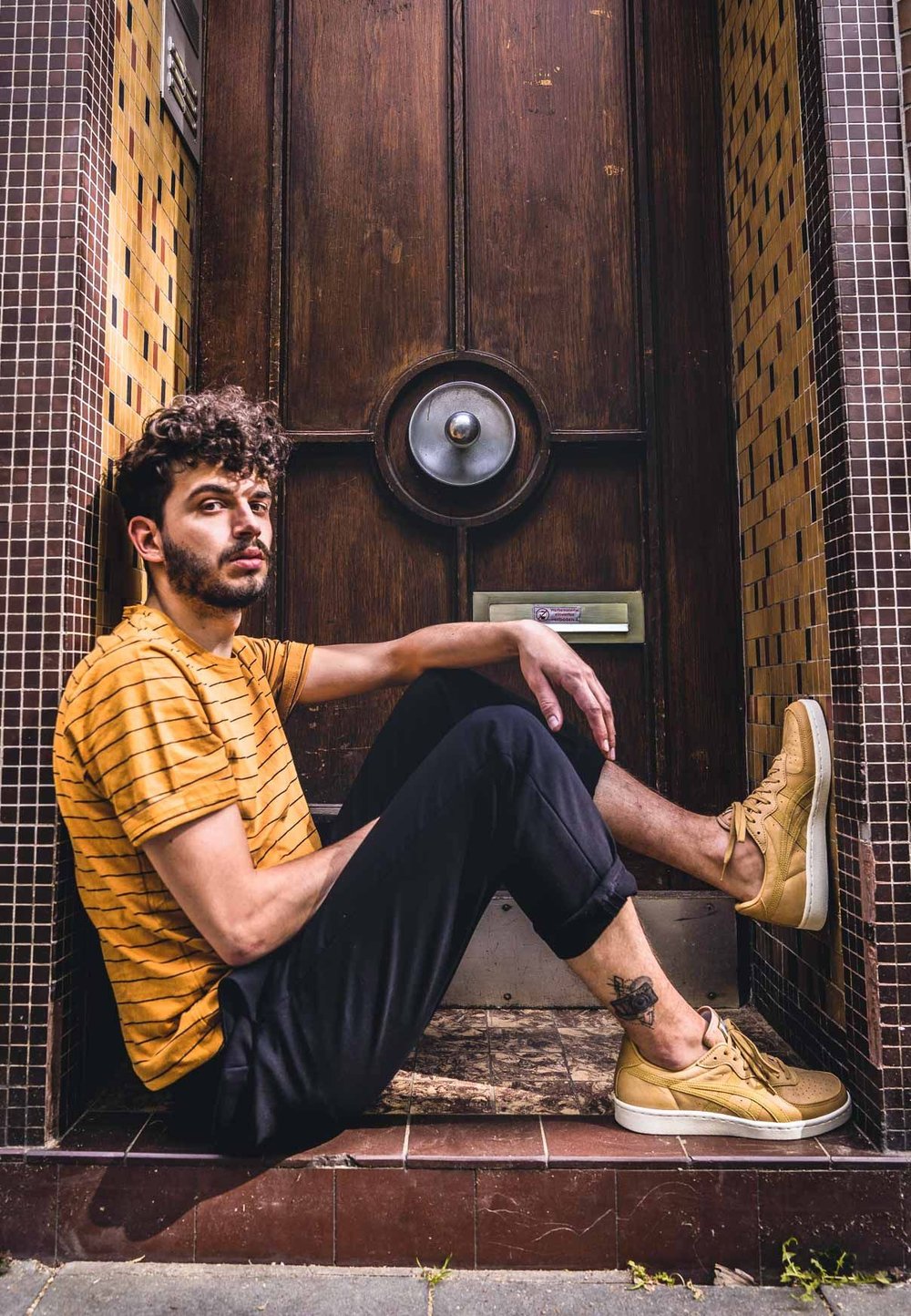 Geel gestreepte overhemd met korte mouwen, zwarte broek en lichtbruine sneakers. Op de achtergrond ziet u een gestructureerde houten deur en een betegelde muur.