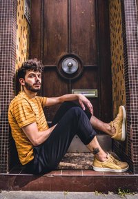 Geel gestreepte overhemd met korte mouwen, zwarte broek en lichtbruine sneakers. Op de achtergrond ziet u een gestructureerde houten deur en een betegelde muur.